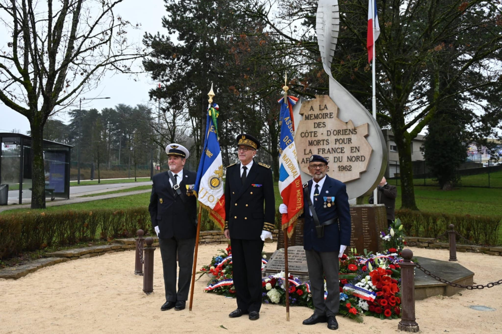 Trois anciens militaires en uniforme se tiennent au garde-à-vous devant un monument aux morts. Deux d’entre eux portent des drapeaux tricolores d’associations, tandis que des gerbes de fleurs rouges, blanches et bleues sont déposées au pied de la stèle. L’inscription rend hommage aux Côte-d’Oriens morts pour la France en Afrique du Nord (1952-1962). Un drapeau français flotte à proximité, dans un cadre arboré et solennel.