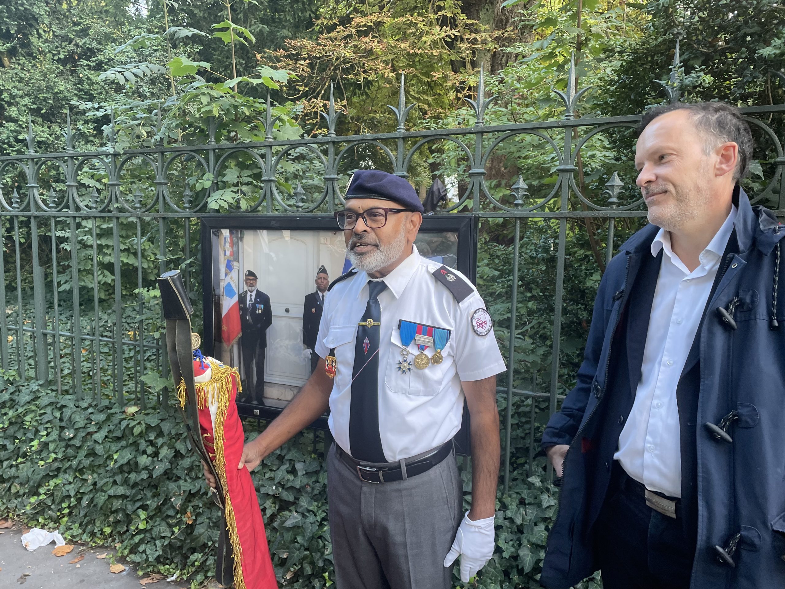Un porte-drapeau en uniforme, décoré de médailles, se tient devant une grille entourée de verdure, accompagné d’un homme en veste sombre.
