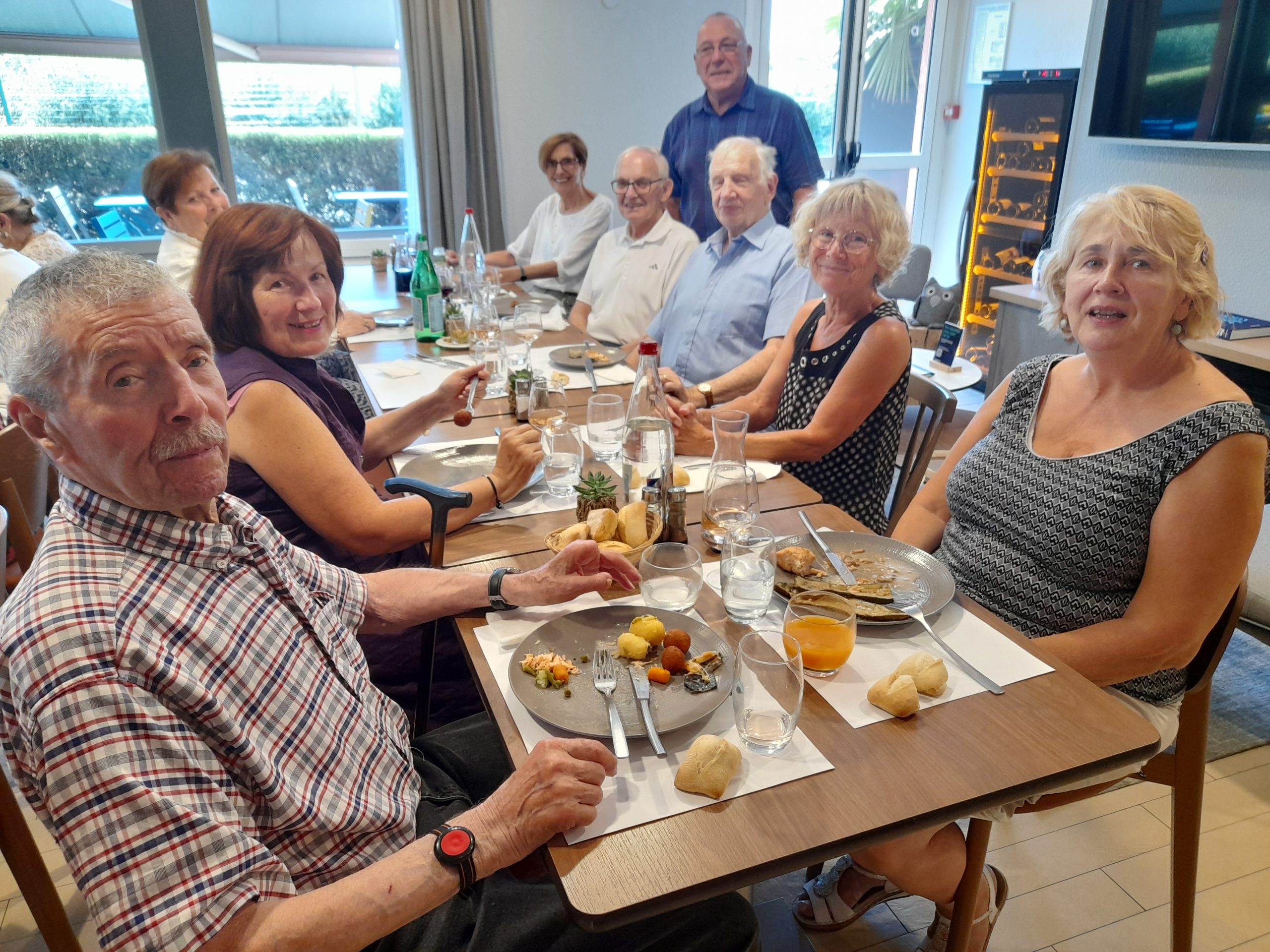 Un groupe de retraités partage un repas convivial autour d’une table dans un restaurant lumineux.