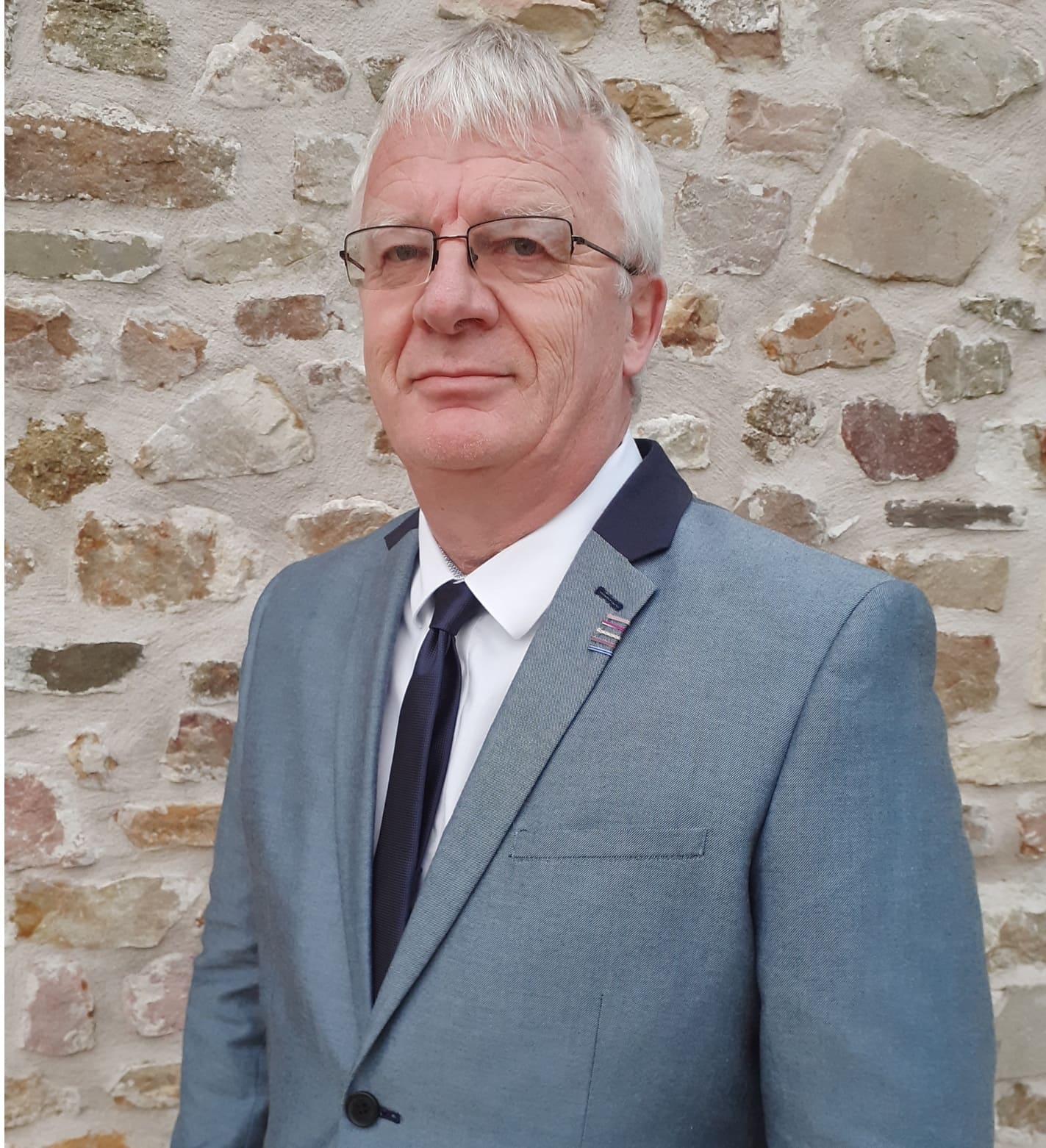 Photo sur fond mur de pierre du président de l'ARM50 posant en costume gris 